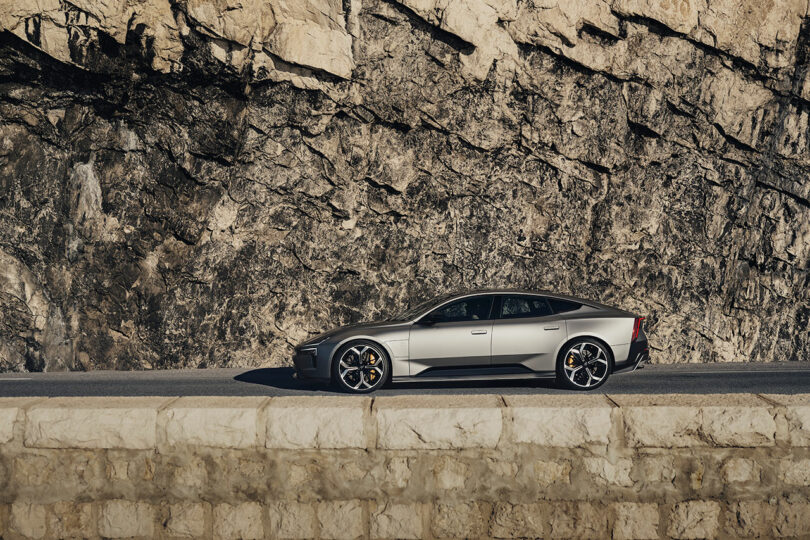 Un sedán deportivo plateado está estacionado en la carretera junto a un acantilado rocoso, el sol proyecta sombras sobre el muro de piedra.