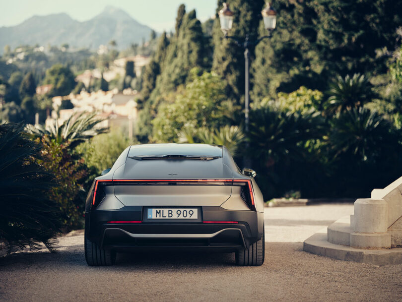Un coche gris mate con matrícula sueca. "Liga Mayor de Béisbol 909" Estacionado en el camino de entrada, rodeado de vegetación y montañas al fondo.