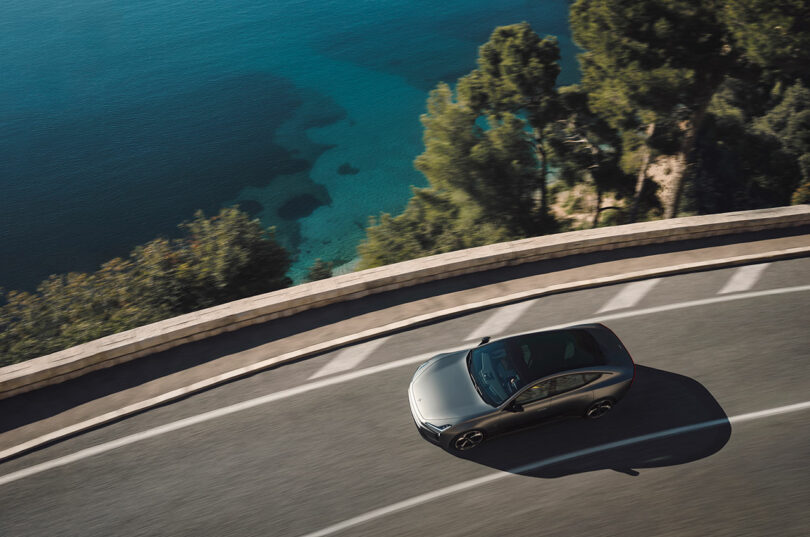 Un coche plateado circula por una carretera costera con curvas, con aguas cristalinas y árboles visibles debajo.