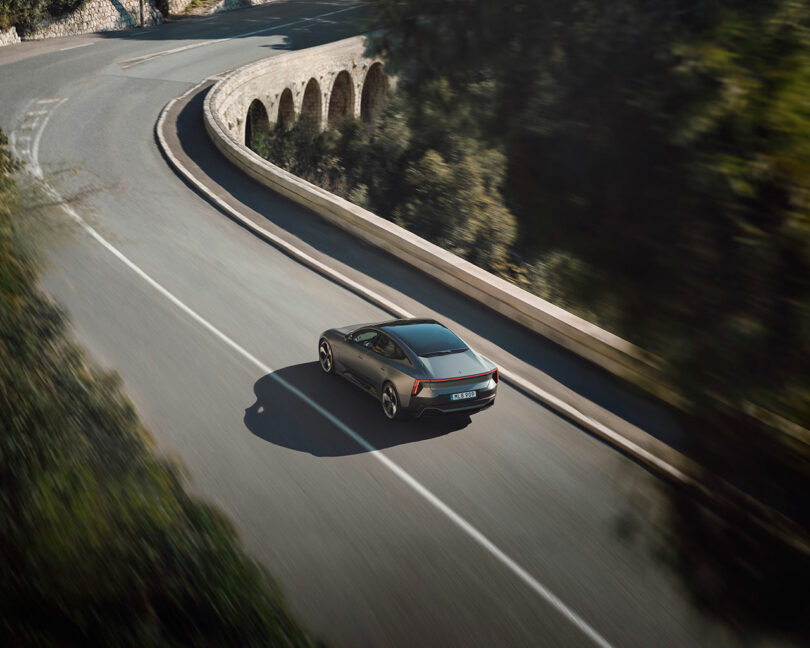 Un coche plateado circulaba solo por una carretera sinuosa junto a un viaducto de piedra, rodeado de árboles verdes.