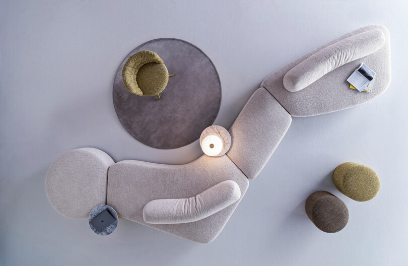 La vista de arriba hacia abajo incluye un moderno sofá modular beige, una alfombra redonda gris, una silla verde, dos taburetes, una lámpara y una mesa auxiliar con revistas y cuadernos.