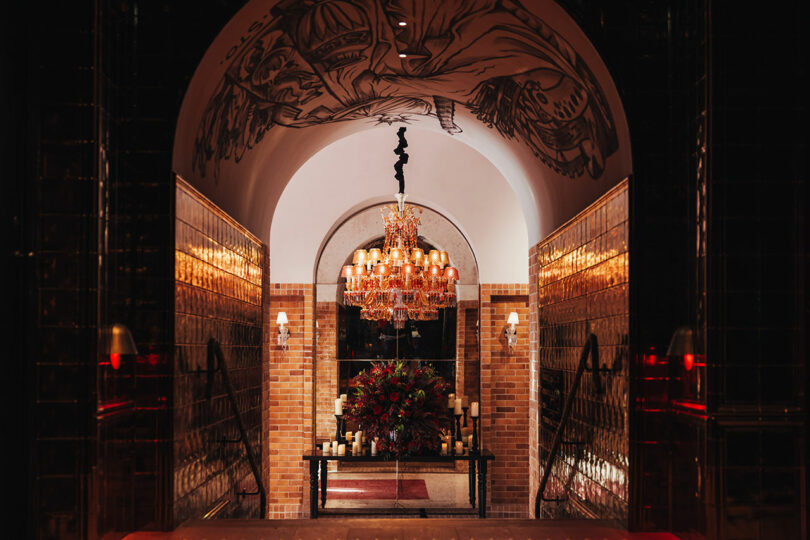 A hallway with brick walls and golden tiles leads to an ornate chandelier, a large floral arrangement, candles, and a mural on the arched ceiling.
