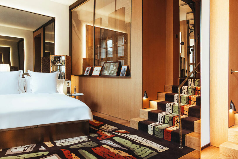 Modern hotel room with a large bed, patterned carpet, wooden walls, and stairs with a colorful runner leading to an upper level.