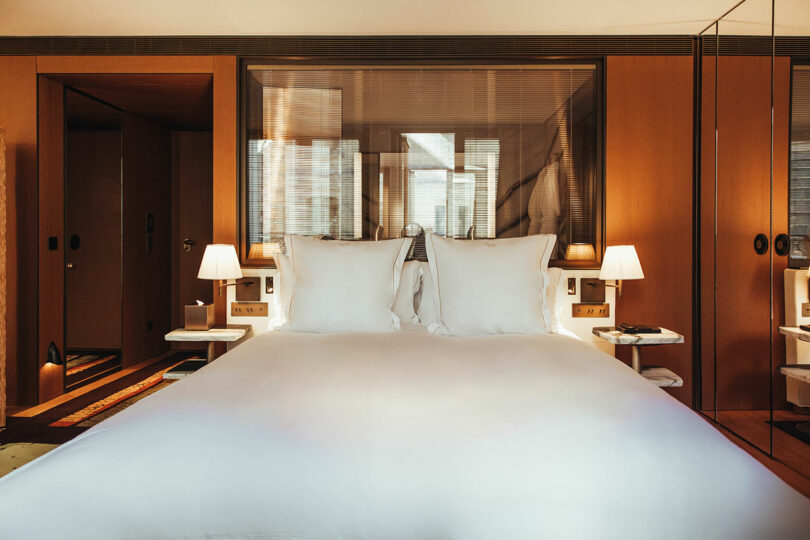 A hotel room with a neatly made double bed, white linens, bedside tables with lamps, wood-paneled walls, and a glass partition behind the bed.