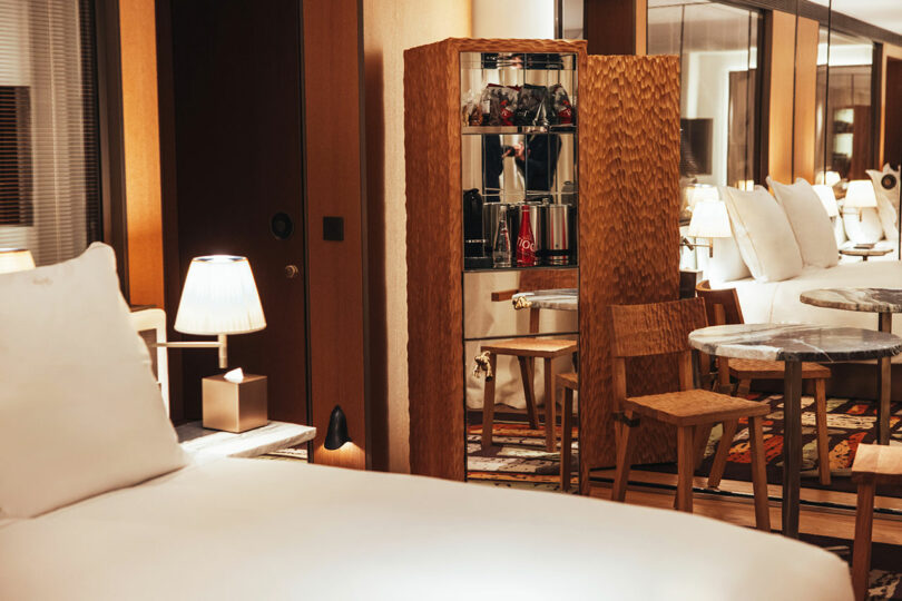 A hotel room with a neatly made bed, a wooden minibar with drinks, two wooden chairs, a small round table, lamps, and a large mirror reflecting the furniture.