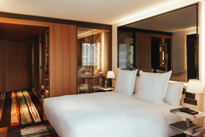 Modern hotel room with a large bed, crisp white linens, bedside tables with lamps, wooden accents, a large mirror, and a patterned rug.