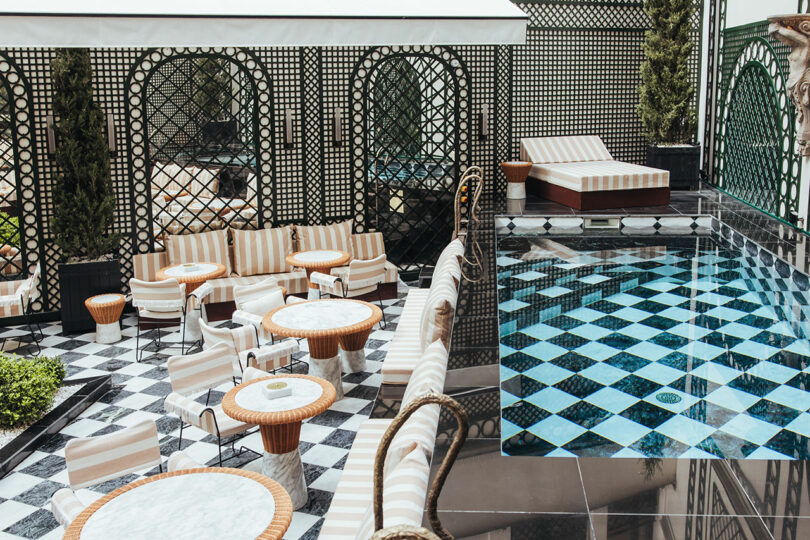 Outdoor seating area with striped chairs and round tables beside a small pool with a black and white checkered tile pattern. Geometric lattice panels surround the space.