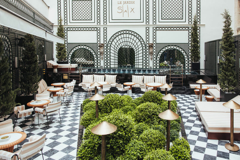 Outdoor courtyard with black-and-white checkered floor, green bushes, striped seating, round tables, and decorative metalwork on the surrounding walls.
