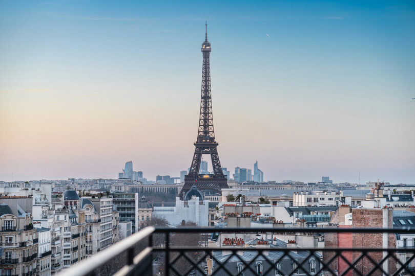 View of the Eiffel Tower rising above Parisian buildings at sunset, with clear skies and cityscape in the background.