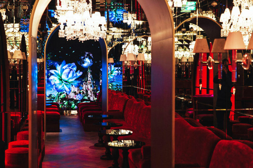 A luxurious lounge with red velvet seating, mirrored walls, chandeliers, and a digital display of large blue flowers at the far end.