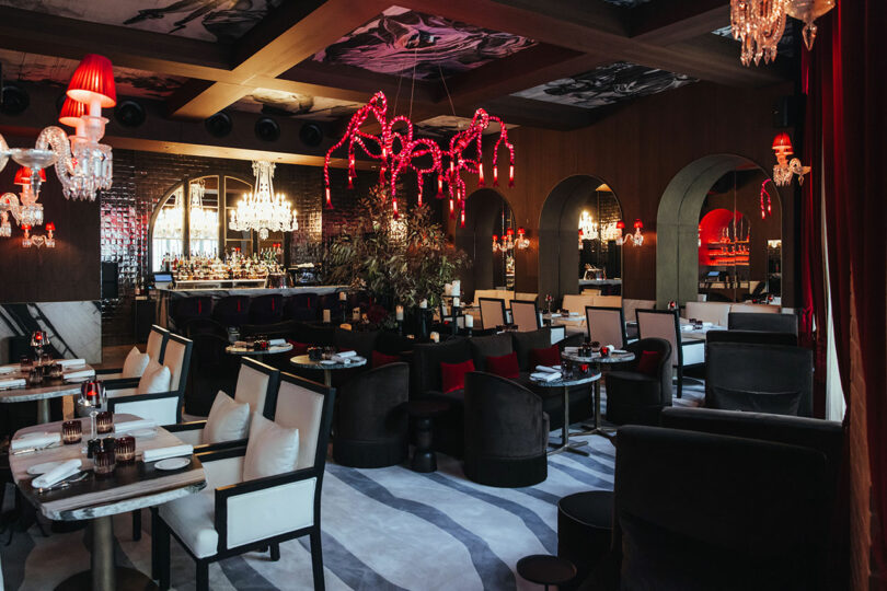 Upscale restaurant interior featuring modern black and white seating, red lighting accents, chandeliers, and arched mirrors along the wall.
