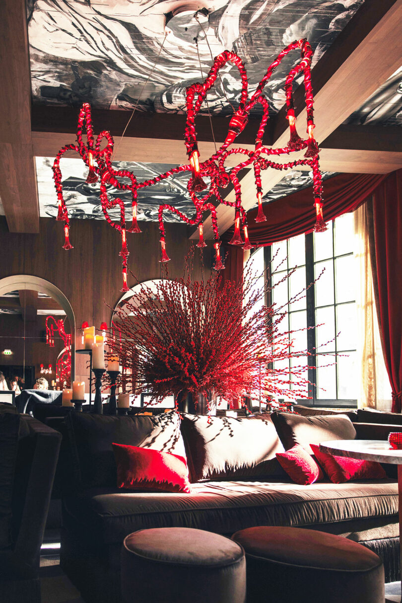 Luxurious lounge with velvet sofas, red accent pillows, large floral arrangement, red chandeliers, and candles; natural light streams through tall windows.