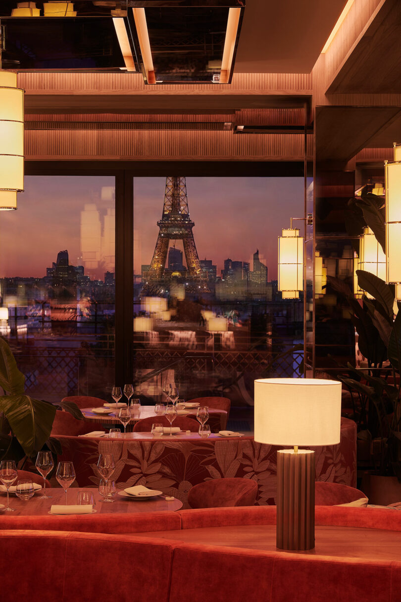 Elegant restaurant interior with red seating and table settings, featuring a large window view of the Eiffel Tower at sunset.