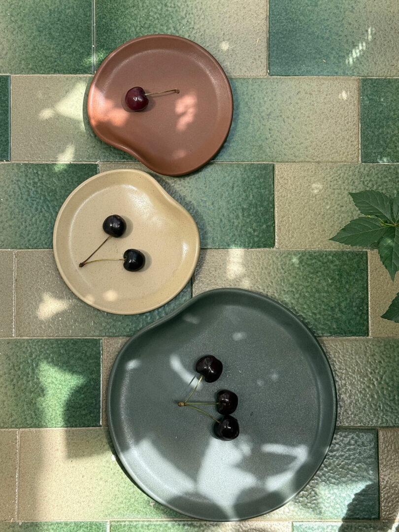 Tres platos de cerámica de diferentes tamaños y colores, cada uno con dos cerezas, se disponen sobre una superficie de azulejos verdes y beige con algunas sombras de hojas.