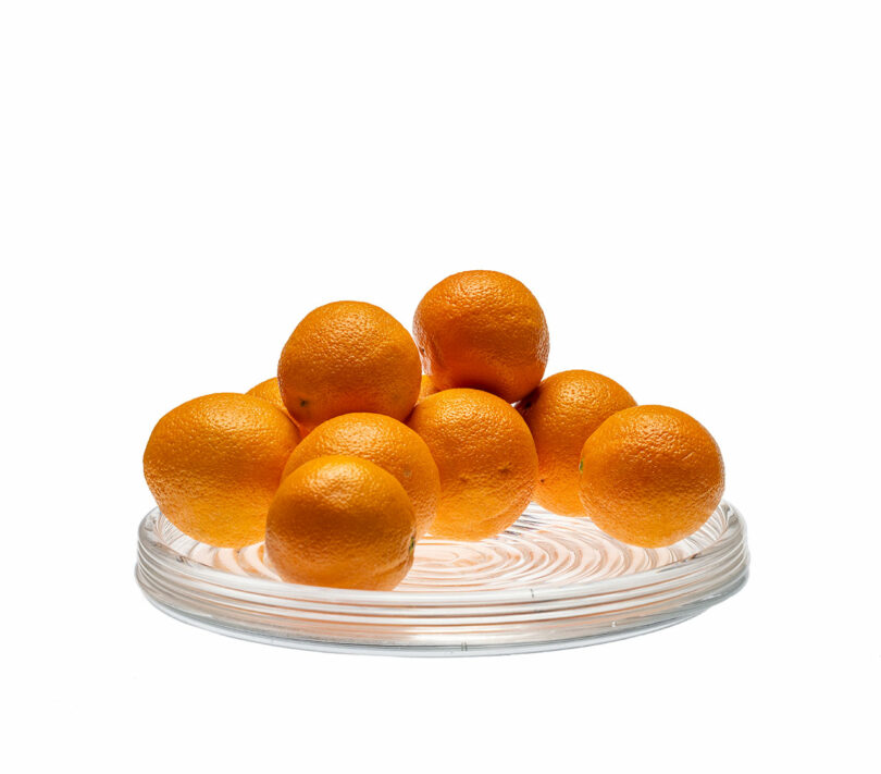 Eight oranges are stacked on a clear glass plate against a white background.