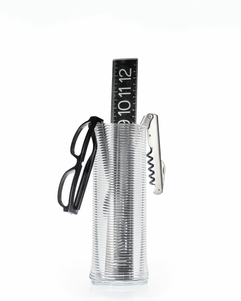 A clear glass holds a black ruler, a pair of black eyeglasses, and a metal corkscrew against a white background.