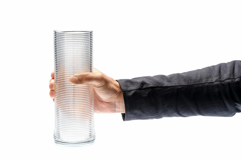 A hand in a dark sleeve holds a tall, empty, ribbed glass cylinder against a plain white background.