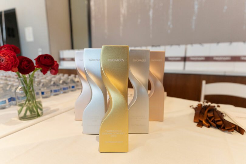 Etiquetas de premio de metal de dos páginas. "IX Concurso de Diseño" Expuesto sobre una mesa decorada con flores rojas, botellas de agua y cintas al fondo.