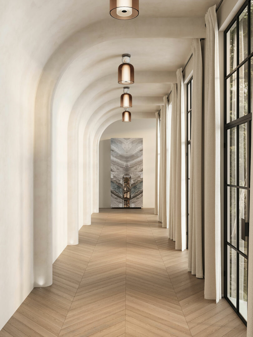 A long hallway with arched ceilings, chevron wood floors, pendant lights, tall windows with beige curtains, and a decorative artwork at the end.
