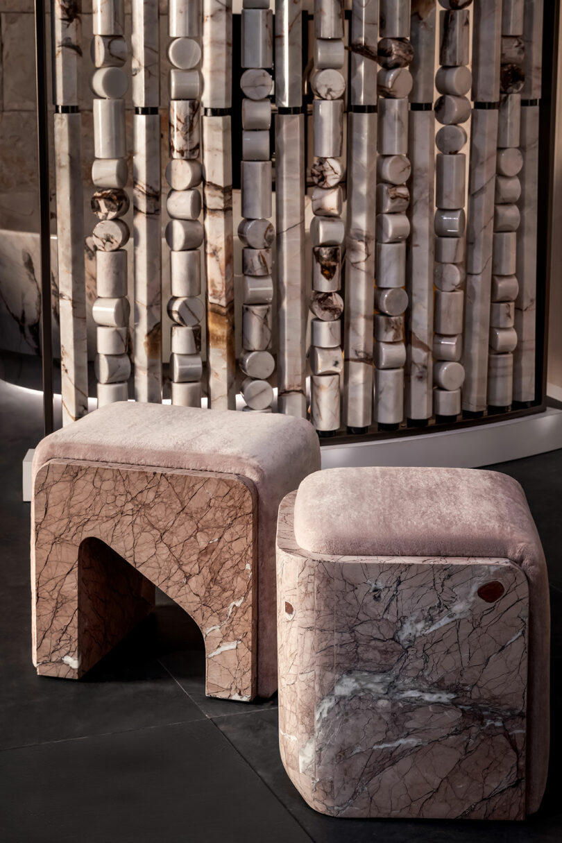 Dos taburetes geométricos de mármol se encuentran en el suelo oscuro, frente a una partición decorativa hecha de bloques de piedra cilíndricos.