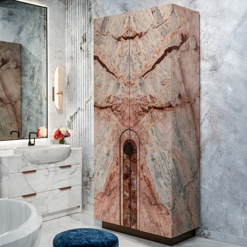 A modern bathroom with marble walls, a white vanity, a round mirror, a stone-patterned cabinet, and a blue textured stool.