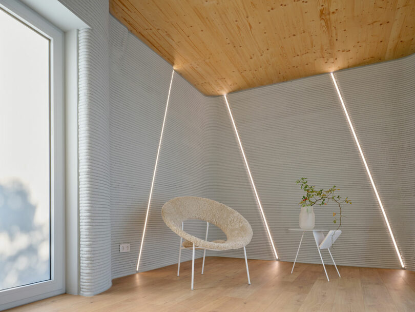 Minimalist room with textured gray walls, wood ceiling and floor, a round beige chair, a small wire table with a white vase and plant, and diagonal light strips on the walls.