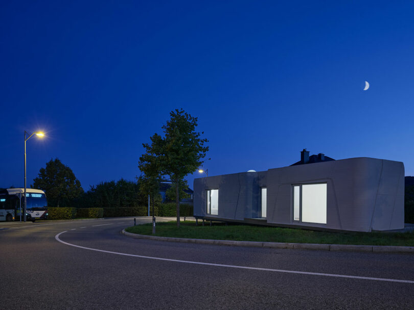 A modern, angular building with illuminated windows sits beside a curved road at dusk, with a streetlight, a tree, and a bus visible under a twilight sky with a crescent moon.