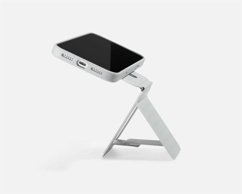A silver smartphone is positioned on a compact, adjustable metal stand against a plain white background.