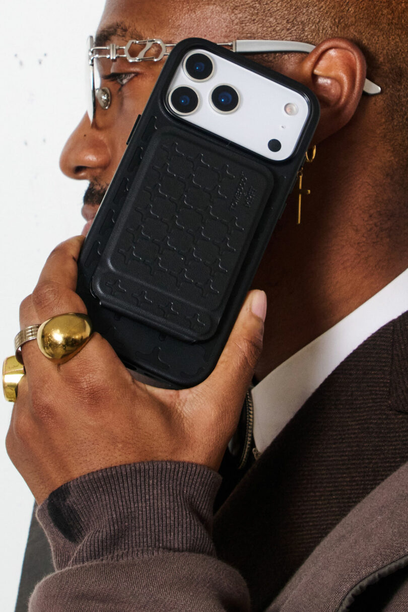 A person holding an iPhone with a rugged black case to their ear, wearing glasses, gold rings, and a dark blazer.