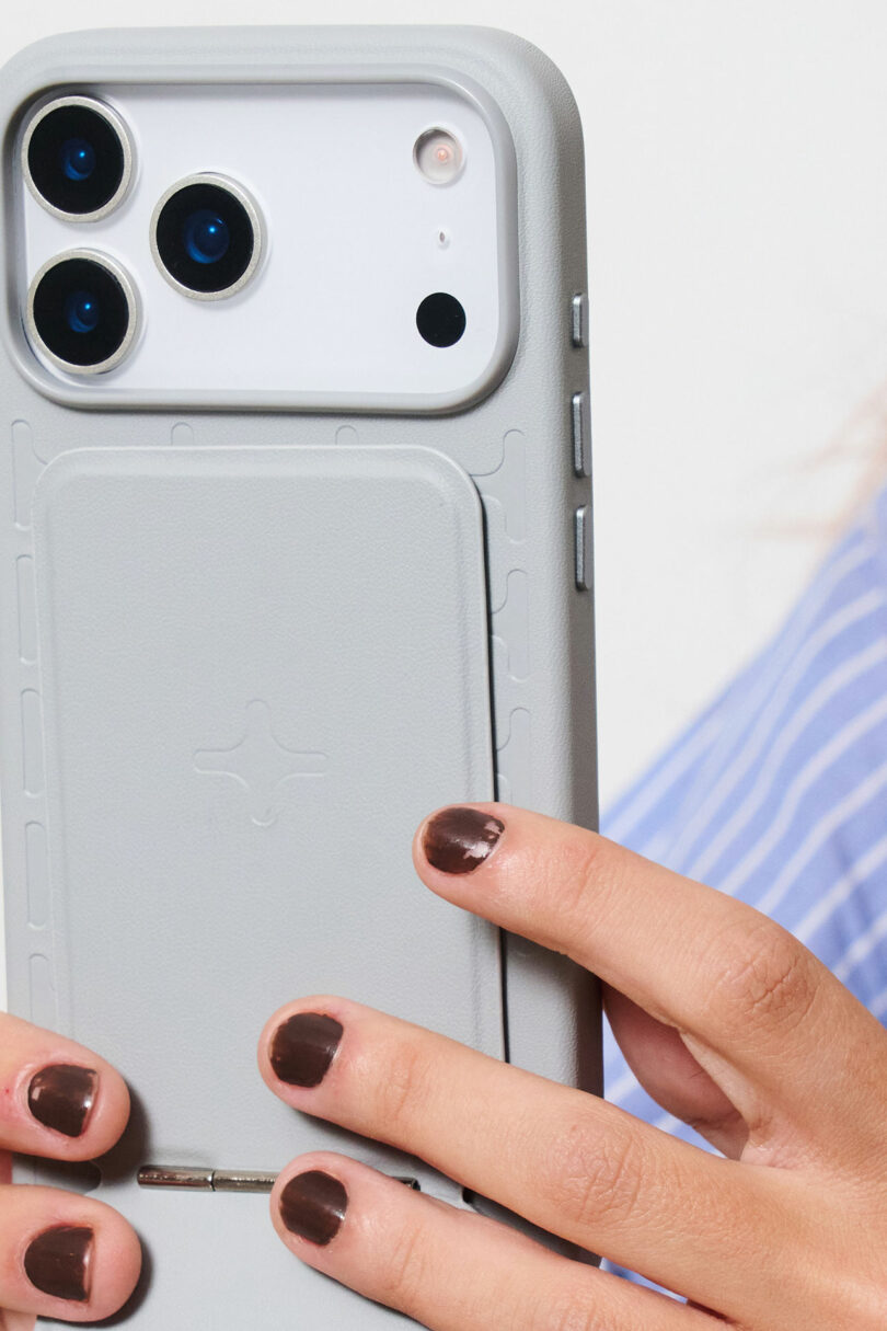A person with dark brown nail polish holds a white smartphone with a gray protective case and card holder attachment.