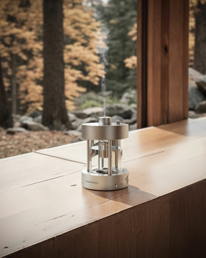 A small, cylindrical aroma diffuser emitting vapor sits on a wooden ledge next to a large window with a view of autumn trees and rocks outside.