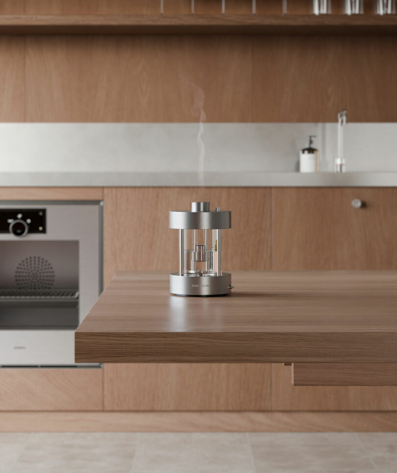 Un moderno difusor de aroma plateado que emite vapor se encuentra en una isla de cocina de madera en una cocina minimalista con gabinetes de madera clara y electrodomésticos de acero inoxidable.