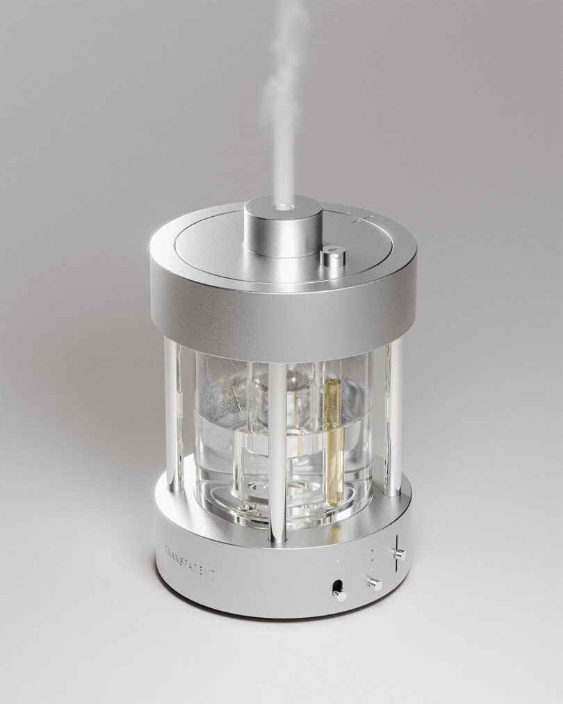 A cylindrical, silver aroma diffuser humidifier emits a thin stream of vapor from the top; its transparent body reveals internal components.