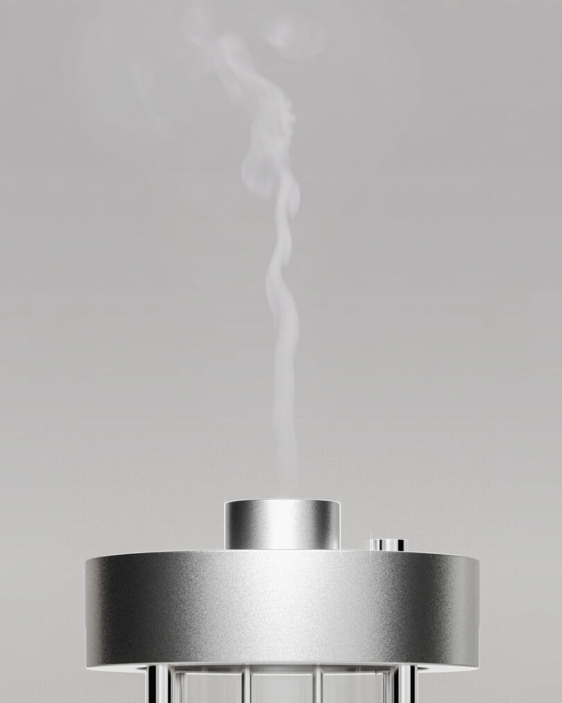A silver cylindrical aroma diffuser emits a thin stream of vapor rising into the air against a plain gray background.