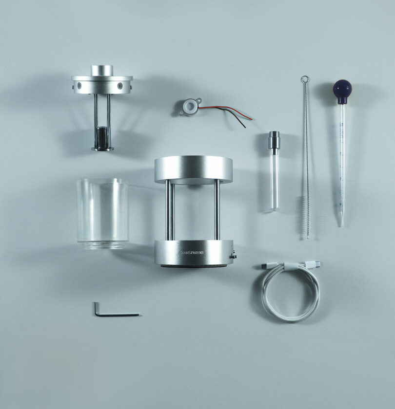 A neatly arranged set of scientific or laboratory equipment, including metal and glass components, a stirrer, a pipette, a beaker, a cable, and a small hex key on a gray background.