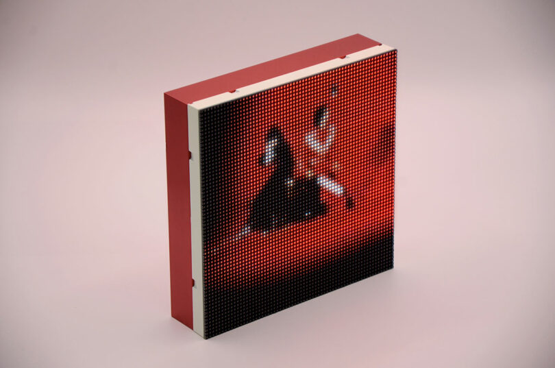 Una pantalla digital con un marco rojo muestra una imagen pixelada de un perro negro sobre un fondo rojo, colocado sobre una superficie lisa de color rosa claro.