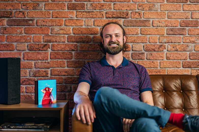 Un hombre barbudo se sienta en un sofá de cuero marrón contra una pared de ladrillos y sonríe a la cámara. Junto a él hay una pequeña estantería con un altavoz y tarjetas o libros de colores.