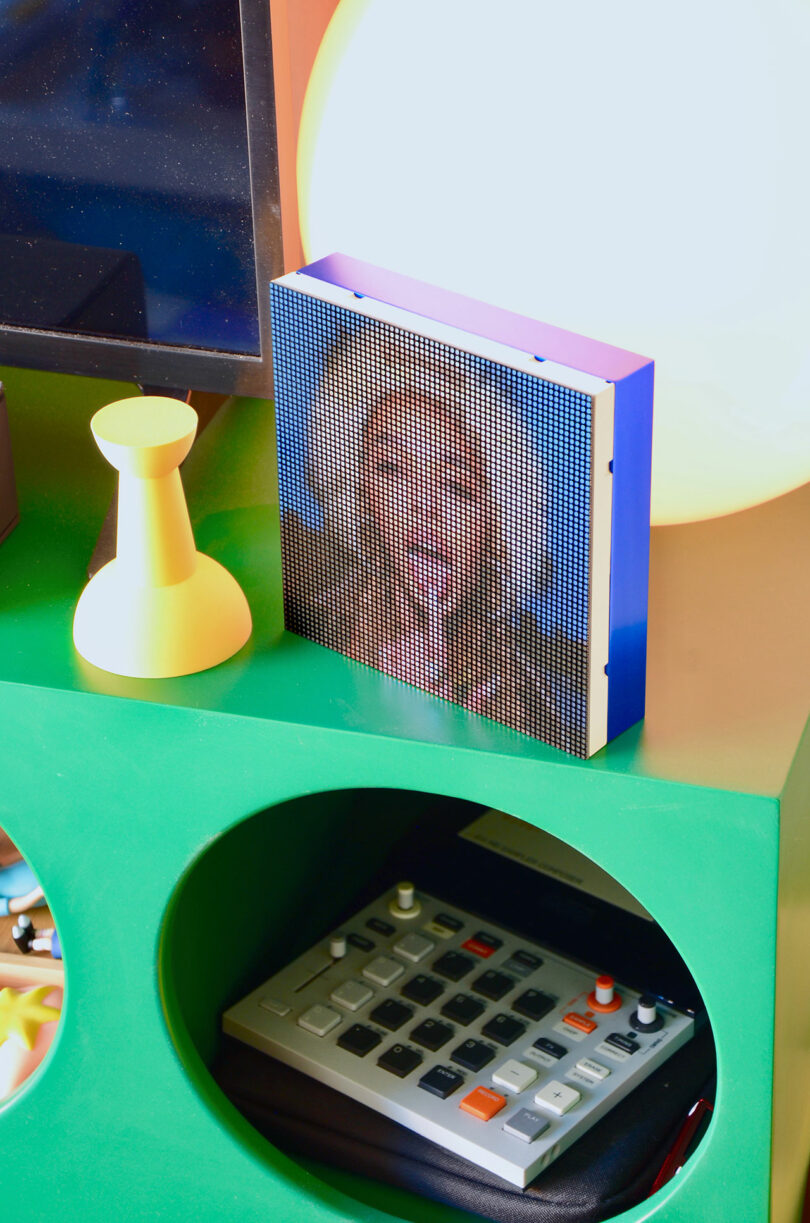 El marco de fotos digital pixelado muestra a una persona sonriente junto a objetos amarillos sobre una mesa verde con una calculadora dentro de un recorte circular.