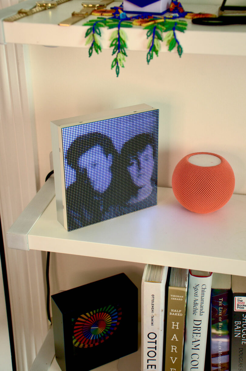 Marco de fotos digital pixelado que muestra dos caras en un estante blanco junto a un altavoz inteligente naranja y varios libros.