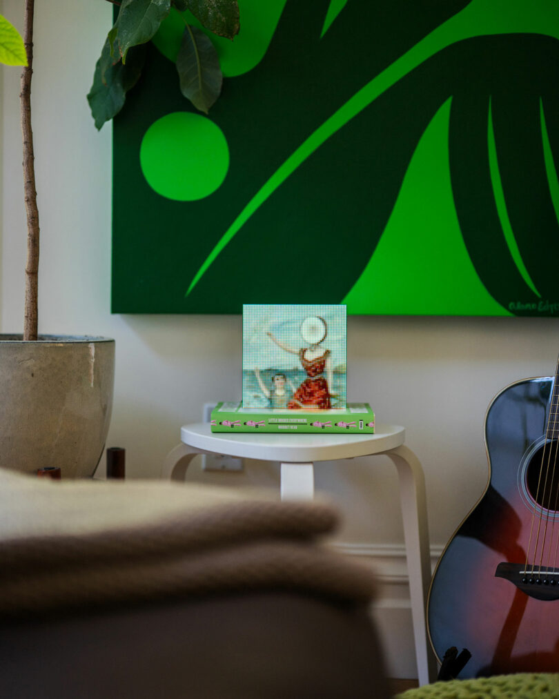 Hay dos libros y un cuadro de pie sobre una pequeña mesa redonda, al lado de una guitarra y plantas, y un gran cuadro abstracto verde cuelga de la pared de atrás.