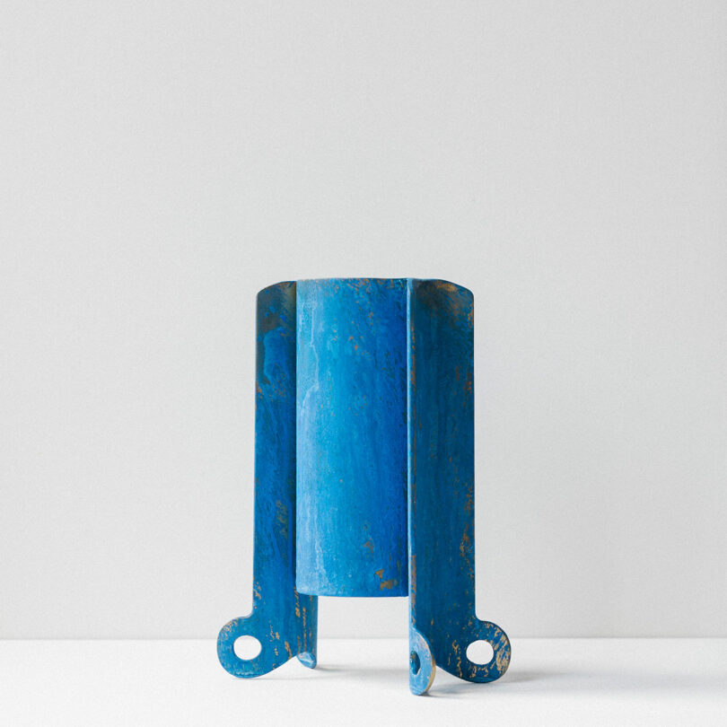 A blue, cylindrical metal object with three curved supports and circular holes at the base, standing upright against a plain white background.