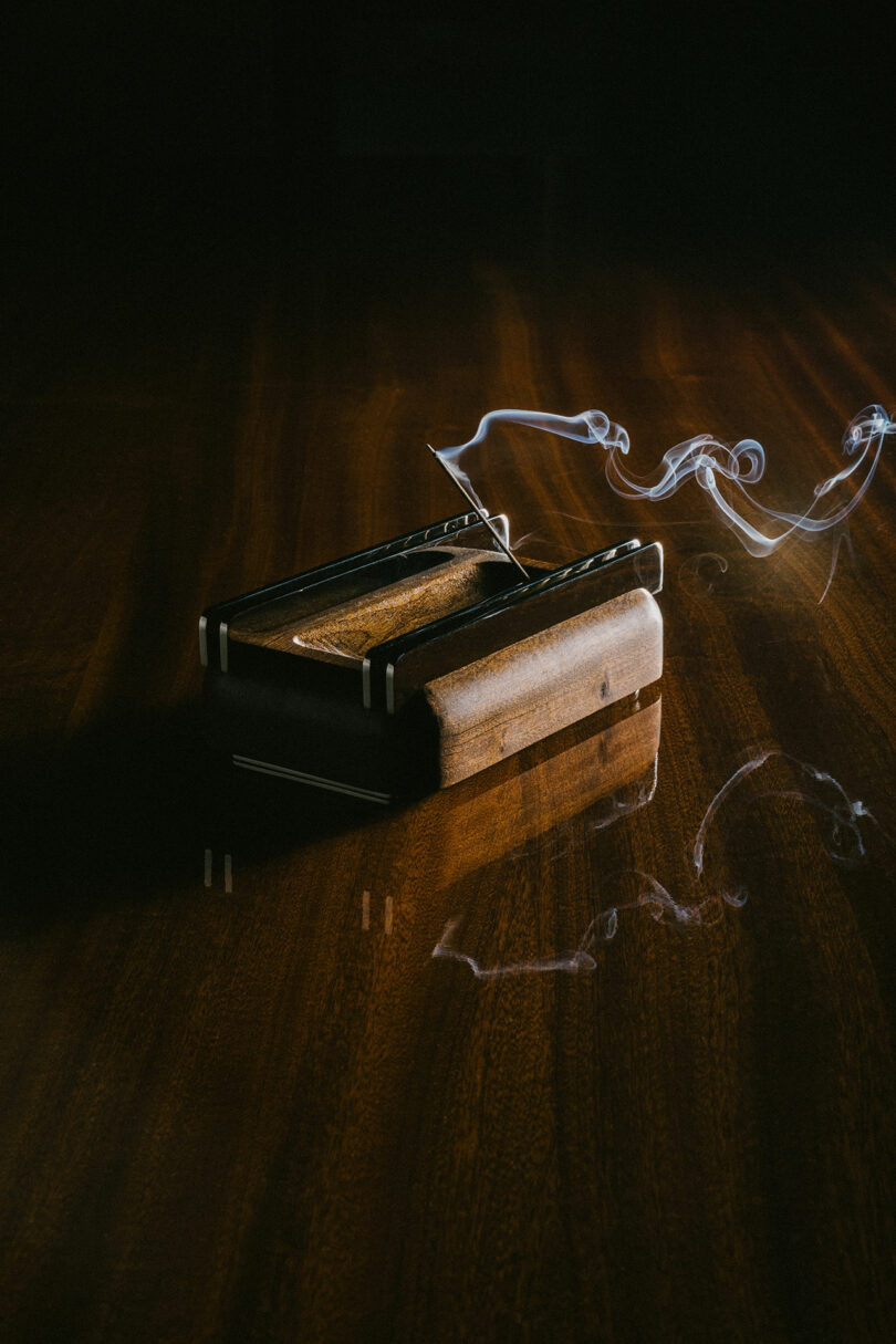 A wooden ashtray with a lit cigarette emitting smoke rests on a polished, reflective wooden surface.