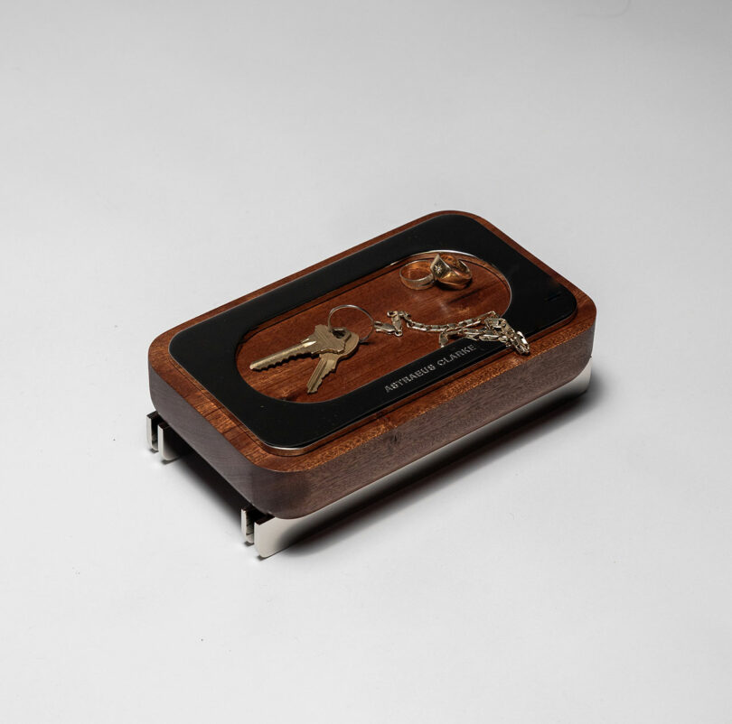 A wooden valet tray with metal legs holds keys, a keychain, and two rings on a white surface.