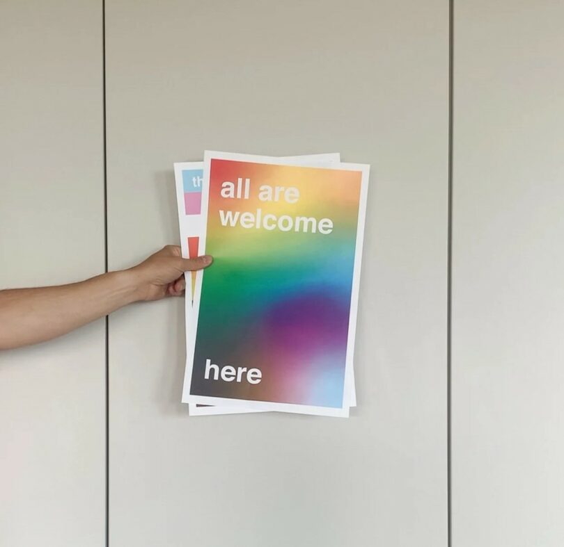 Una mano sostiene un cartel que muestra texto y fondo degradado de arco iris "bienvenidos a todos aquí