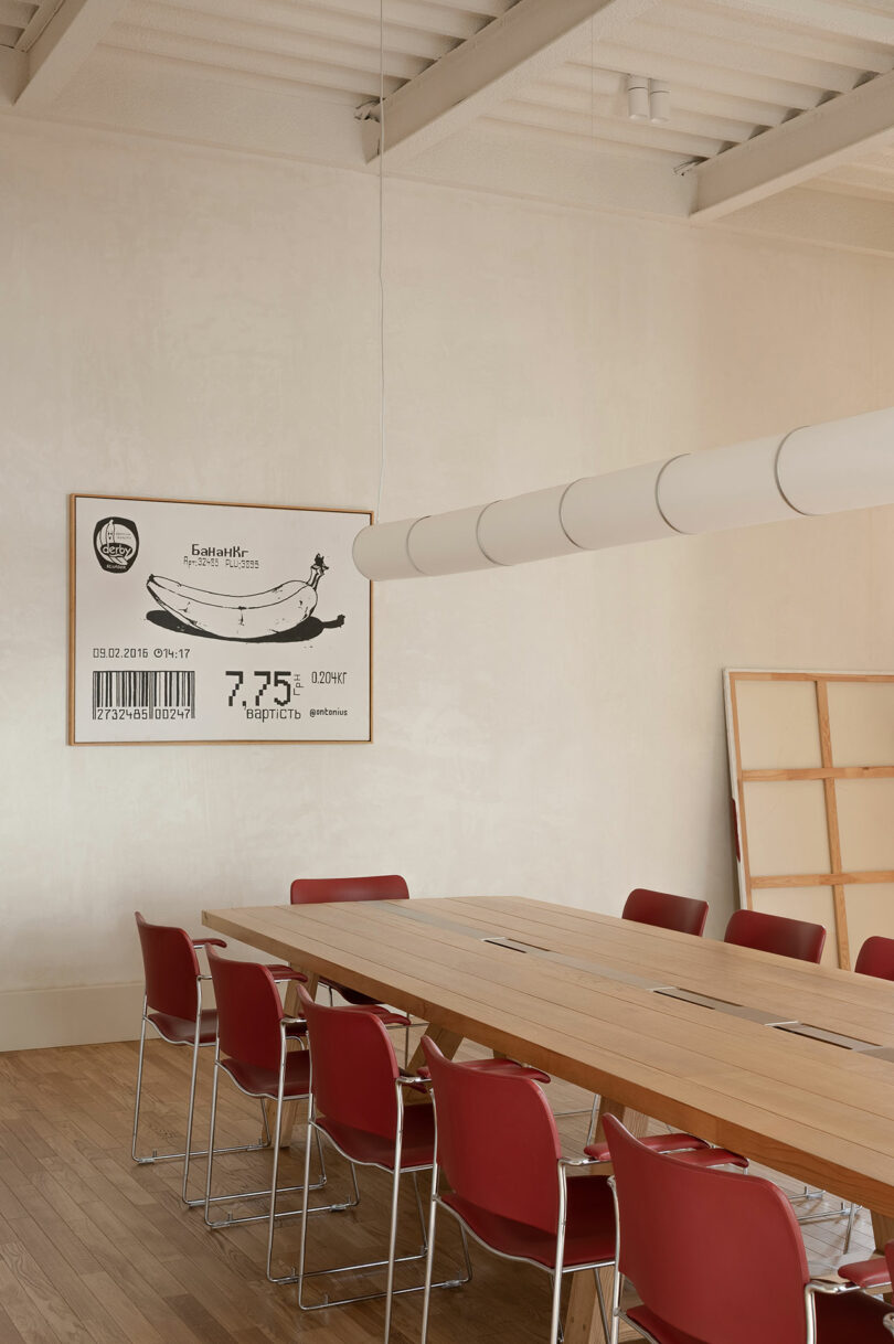 Conference room with a long wooden table, red chairs, and a large wall art resembling a banana with a barcode and numbers, set against light-colored walls and ceiling.