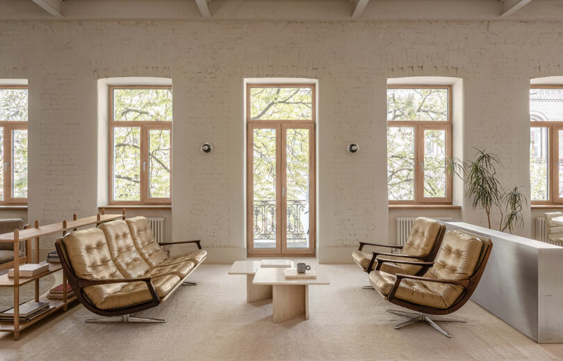 A modern living room with two beige sofas, two armchairs, a coffee table, and large windows letting in natural light. A potted plant is in the corner.