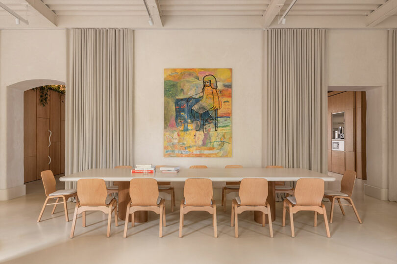 A long dining table with ten wooden chairs, several books on the table, and a colorful abstract painting on the wall in a neutral-toned room with curtains and archways.