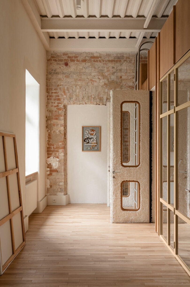 A bright hallway with exposed brick and plaster walls, a textured glass door, wooden floor, and a framed artwork that reads "Ping Pong Pitch".