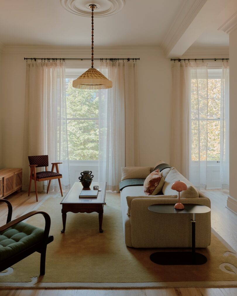 Salón con grandes ventanales, cortinas transparentes, sofá, sillas de madera, mesa de centro y alfombra. La luz natural ilumina el espacio.