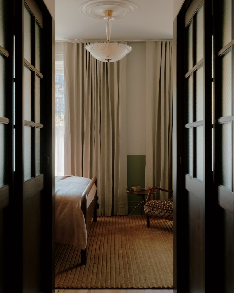 Un dormitorio pequeño con una cama de madera, ropa de cama de color beige, un sillón, cortinas de suelo a techo, una alfombra de rayas y lámparas de techo.